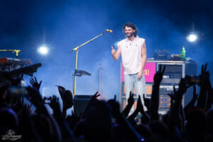 Male singer on stage giving a thumbs-up, wearing a white sleeveless shirt and light jeans, with blue stage lights and crowd raising hands in the foreground.