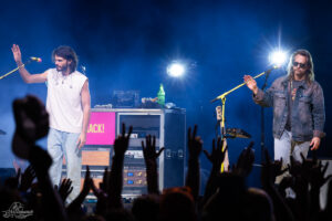 Two musicians perform on a smoky stage under blue lights; one in a white sleeveless shirt raises a hand, the other in a denim jacket and sunglasses sings into a mic, crowd raises hands in front.