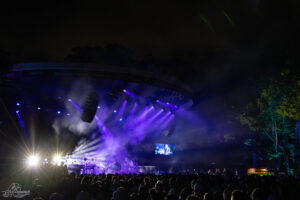 Outdoor night concert with a large crowd watching a stage lit in purple beams and smoke, trees in the background.