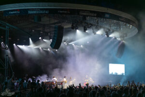 Open-air concert stage with a band performing amid bright spotlights and smoky atmosphere, crowd raising hands in the foreground.