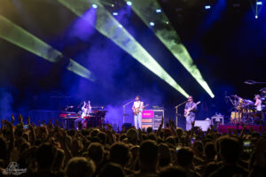Live concert scene with a band on stage, guitarist center, bassist to the right, drummer on the far right, and a cheering crowd under colorful spotlights.