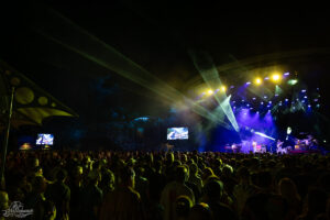 Outdoor nighttime concert with a large crowd facing a brightly lit stage, colorful spotlights, and screens showing performers in the distance.