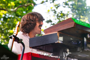 Male musician with shaggy hair and moustache singing into a keyboard rig outdoors, with a guitar nearby.