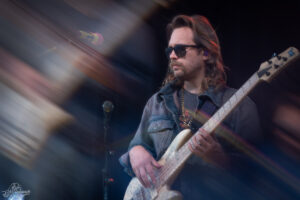 Bassist wearing sunglasses and a denim shirt plays a light-colored bass on stage with colorful light trails in the background.