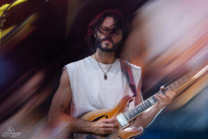 Guitarist with long dark hair wearing a white sleeveless shirt, playing an orange electric guitar on stage with a colorful strap and motion-blurred background.