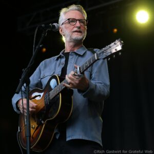 Guitarist with gray hair and glasses sings into a microphone on stage while playing an acoustic guitar.