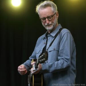 Man with gray hair and beard wearing glasses plays an electric guitar on stage, wearing a blue shirt and black strap with warm spotlights in the background.