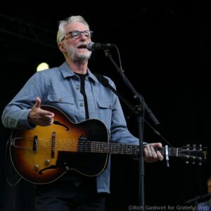 Male musician singing into a microphone while playing a sunburst acoustic guitar on stage, wearing glasses and a blue denim shirt.