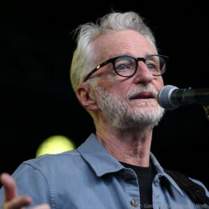 Close-up of an older man with gray hair and glasses speaking into a microphone on stage