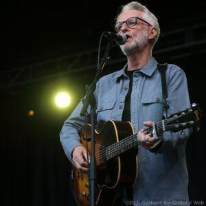 Male guitarist singing into a microphone on stage, wearing glasses and a blue shirt.