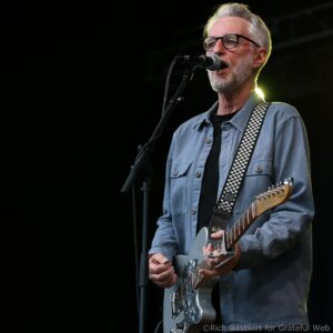 Male guitarist with gray hair and beard singing into a microphone while playing a silver electric guitar wearing a blue denim shirt and a black-and-white checkered strap.