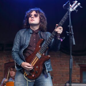 Male guitarist playing a brown bass guitar on stage, wearing a denim jacket and red patterned shirt against a dark backdrop.