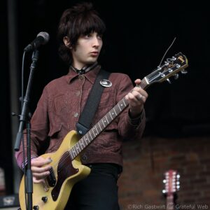 Male guitarist performing on stage, wearing a brown corduroy jacket and playing a cream-colored electric guitar beside a microphone stand.