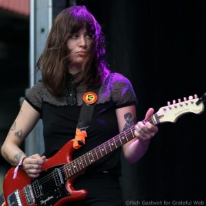 Guitarist playing a red electric guitar, seen from the torso up, black outfit with dotted mesh top and visible tattoos on arms under stage lighting with a purple hue.