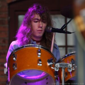 Female musician singing into a microphone with purple stage lighting, drums visible in the foreground.