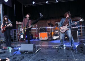 Wide shot of a rock band performing on an outdoor stage with three guitarists and a drummer in the background against a brick wall backdrop.