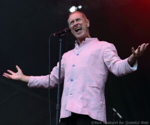 Male singer on stage, mouth open in a powerful note, gripping a microphone with outstretched arms in a pink shirt.