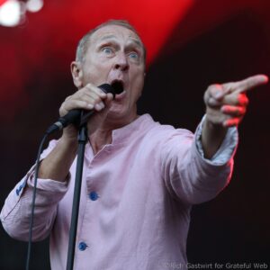 Male singer on stage, pink shirt, holding microphone and reaching toward the audience with a red-lit background.