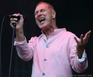 Male singer wearing a pale pink shirt passionately singing into a handheld microphone on stage.