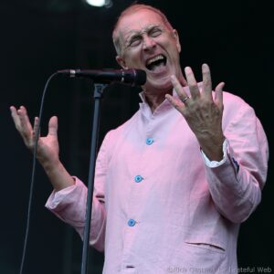A man singing into a microphone on stage, wearing a light pink shirt and expressive with closed eyes and open mouth.