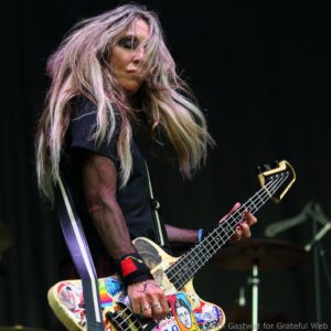 Female guitarist playing a sticker-covered bass on stage, with long, windblown hair partially obscuring her face.
