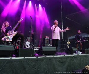 Live onstage performance with guitarist left, singer center in pale shirt, under purple stage lights.