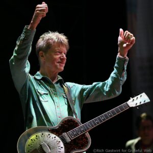 A musician with both arms raised in the air, playing a metal‑bodied resonator guitar against a dark stage background.
