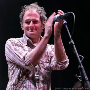 Smiling male musician on stage in a plaid shirt, with a decorative guitar strap and microphone nearby, mid-performance.