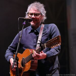 Male guitarist singing into a microphone on stage, wearing glasses and a dark shirt.