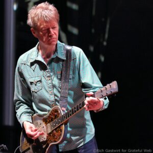 Male guitarist with blond hair wearing a turquoise patterned shirt, playing a resonator guitar on stage.