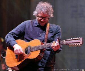 Male musician wearing glasses and a blue shirt plays a wooden acoustic guitar on stage with a patterned strap visible in the foreground on stage lights. Pastel blur in background, highlighting performance stance.