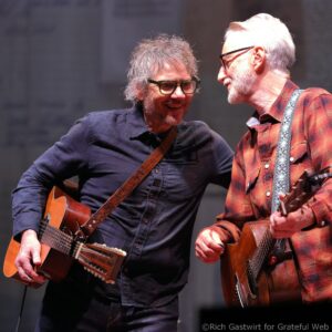 Two male guitarists on stage sharing a moment, each holding an acoustic guitar and facing each other with smiles on a live performance.