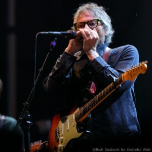 Male guitarist with green-rimmed glasses singing into a microphone while playing a sunburst electric guitar on stage.