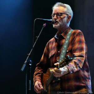 Male musician in a plaid shirt wearing glasses, singing into a microphone while playing an acoustic guitar on stage.