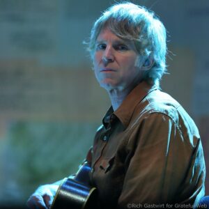 Close-up of a person with short blond hair under blue stage lighting, wearing a brown shirt and holding an acoustic guitar across their lap.
