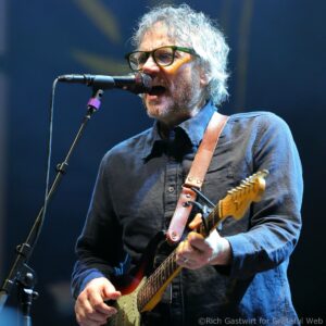 Male musician with gray hair and beard, wearing green-framed glasses, singing into a microphone while playing a sunburst electric guitar on stage.