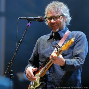 Male guitarist with gray hair and green glasses, singing into a microphone while playing a cream-colored electric guitar on stage.