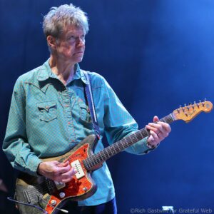 Male guitarist with gray hair wearing a teal patterned shirt playing a sunburst electric guitar on a blue-lit stage.