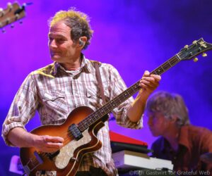 Male guitarist in a plaid shirt playing a sunburst electric guitar on a stage bathed in purple lighting.