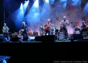 Band performing on a stage bathed in blue spotlight with guitars and microphones and a postered backdrop.