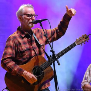 Male musician with glasses singing into a microphone while playing an acoustic guitar on a stage with purple lighting.