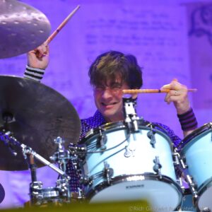 Drummer mid-performance holding drumsticks above a blue-and-silver kit on a purple-lit stage.