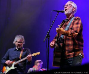Male guitarist and singer with gray hair and beard, wearing glasses and a red plaid shirt, performing on stage.