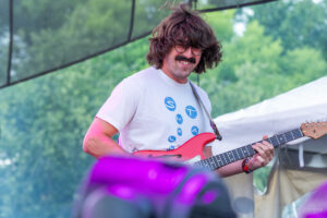 Man with shoulder-length hair and mustache plays a red electric guitar on an outdoor stage, wearing a white graphic T-shirt.