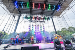 Outdoor concert stage with five musicians performing under a bright multicolor light rig.