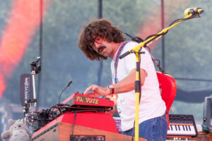Musician playing a red keyboard on stage, wearing a white t-shirt and jeans, with a yellow microphone stand nearby.