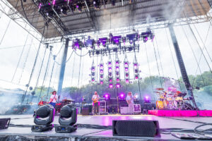 Live band performing on an outdoor festival stage with purple lighting and smoke, guitarist center, drummer right.