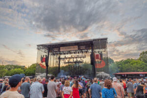 Outdoor concert crowd facing a large stage framed by metal rigging, with banners and smoke as the sun sets behind the venue.