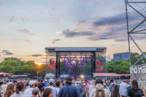 Outdoor concert at sunset with a large crowd facing a brightly lit stage and purple lights behind the performers.
