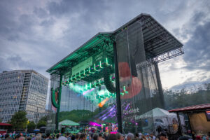 Outdoor concert stage at Red Hat Amphitheater with green and purple lights illuminating performers; crowd gathered in front under a cloudy sky.